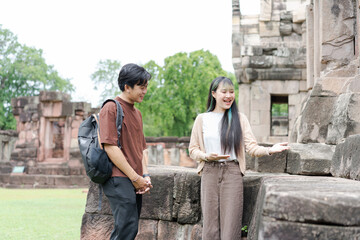 Fototapeta premium Happy Asian young man and woman tourist couple with backpacks holding smartphone, smiling while exploring and sightseeing together at historical ancient stone castle site during summer vacation.