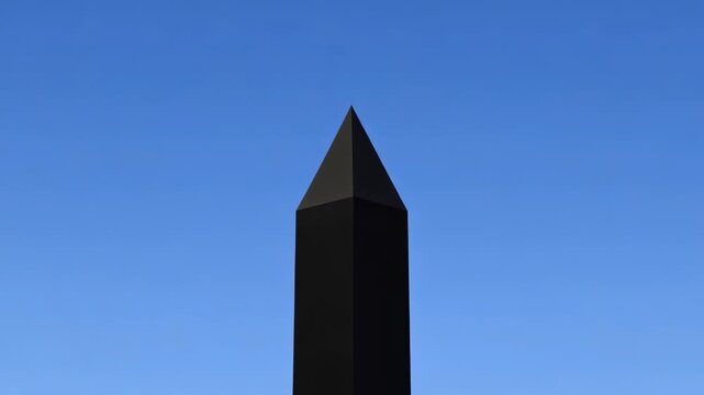 Dark monolithic obelisks stand against a clear blue sky with one exploding in bright white energy