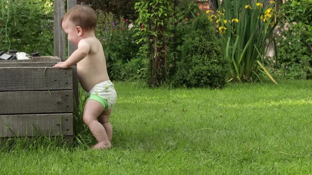 A toddler stands near a flower bed, exploring its contents. He picks up clumps of earth and grass with his hand and throws them down next to him. The toddler really enjoys this activity