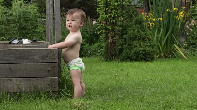 A toddler stands near a flower bed, exploring its contents. He picks up clumps of earth and grass with his hand and throws them down next to him. The toddler really enjoys this activity