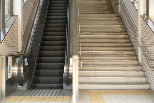点字ブロックが敷かれた駅構内のエスカレーターと階段の構図。非常停止ボタンと安全警告ステッカー