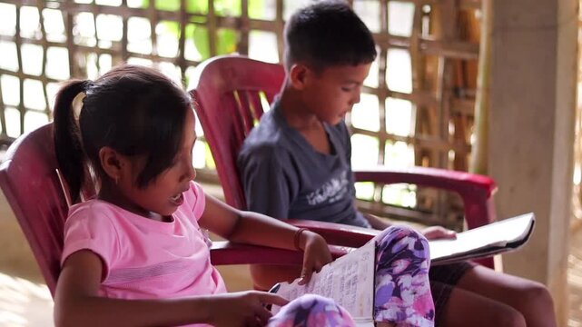 Two children practice a song together while following lyrics in notebooks inside a simple rural home. The warm indoor scene highlights childhood, music education, singing practice.