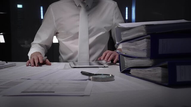 Businessman working overtime in a dark office, holding a tablet and analyzing financial documents, piles of folders and paperwork on the desk showing dedication and hard work