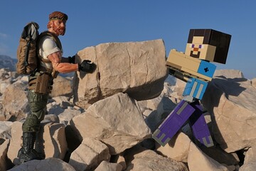 Obraz premium Minecraft action figure of Steve with GI JOE action figure of survival instructor Outback, moving large square shaped coastal rock across the other on Croatian beach, blue summer skies in background. 