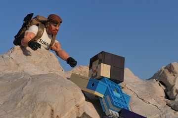 Obraz premium GI JOE action figure of survival instructor Outback, from Classified series, reaching Minecraft Steve action figure to help him climb on coastal rocks, clear blue skies in background, evening sunshine
