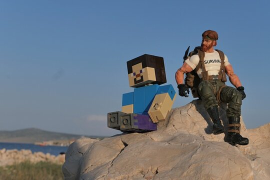 Action figure of Minecraft hero Steve and GI JOE Classified series survival instructor Outback sitting on coastal rock in Croatia, sunlit by evening sunshine, blue skies in background.