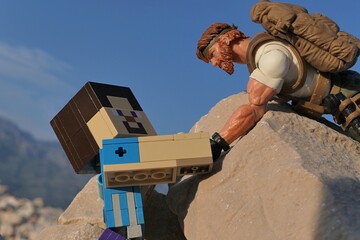 Obraz premium GI JOE action figure of survival instructor Outback, Classified series, helping figure of Minecraft Steve to climb on coastal rock in Croatia. Summer clear blue skies. 