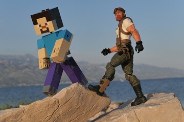 Obraz premium Figures of Minecraft game hero Steve and GI JOE Classified edition survival instructor Outback trekking on sharp coastal rock of Croatian shoreline, bay in Razanac and Velebit mountain in background. 