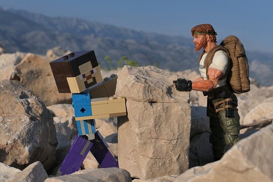 Action figure of Minecraft Steve and GI JOE Classified edtition survival instructor Outback lifting large square block on on Croatian beach, summer daylight sunshine. 