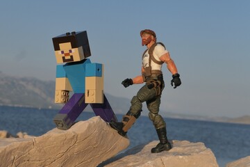 Obraz premium Figures of Minecraft game hero Steve and GI JOE Classified edition survival instructor Outback walking on sharp coastal rock of Croatian shoreline, bay in Razanac and Velebit mountain in background. 