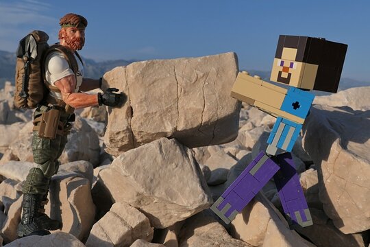 Action figures Minecraft Steve and GI JOE Classified edtition survival instructor Outback placing large square block on on Croatian beach, summer daylight sunshine. 