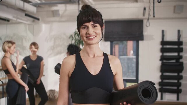 Cheerful yoga instructor smiling at camera. Women standing with mats in meditation studio. Women in sportswear preparing for yoga class. Smiling woman standing foreground and holding mat.
