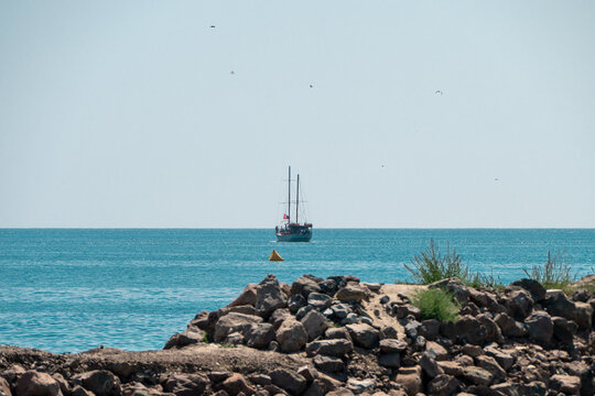 a tranquil seascape featuring a distant two-masted sailing ship navigating a calm blue ocean, framed by a rugged, rocky breakwater in the foreground.
