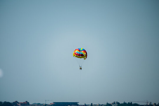 a brightly colored parasail gliding high in a clear blue sky, with the distant, dark silhouette of a coastal cityscape visible along the horizon below.