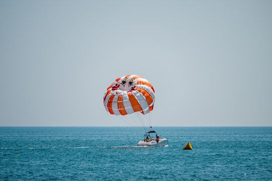a bright orange and white striped parasail gliding above a vibrant blue ocean, towed by a small white motorboat under a clear, cloudless sky.