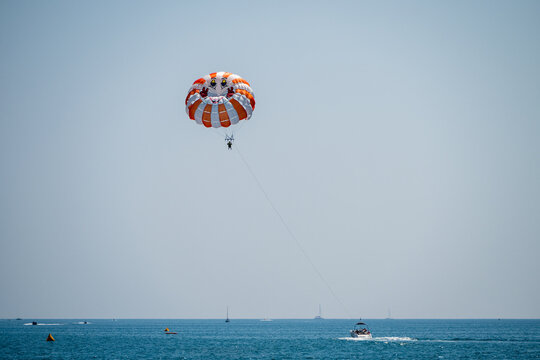 a bright orange and white striped parasail flying high against a clear blue sky, tethered by a long line to a distant motorboat speeding across the vast ocean.