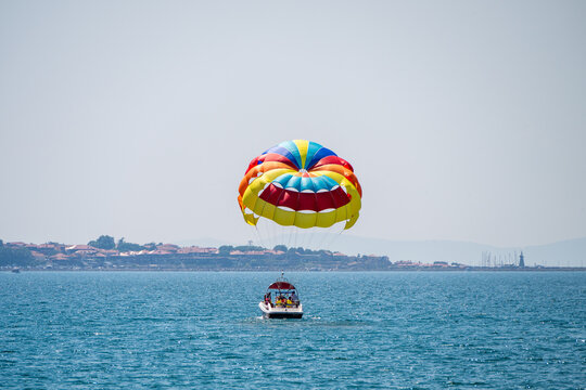 a vibrant, multi-colored parasail being towed by a motorboat across a sparkling blue sea, with a picturesque coastal town visible along the distant shoreline under a clear sky.