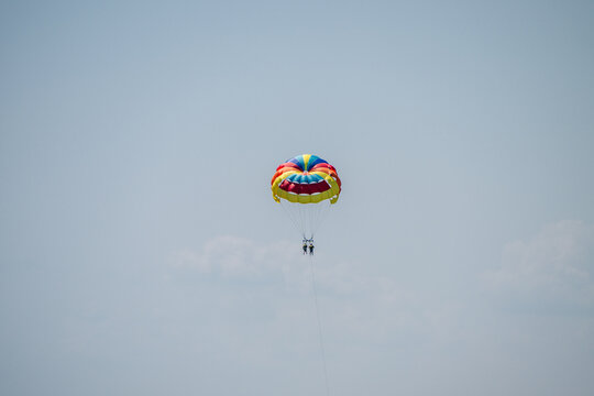 two people enjoying a tandem parasailing flight under a brightly colored parachute against a vast, minimalist blue sky.