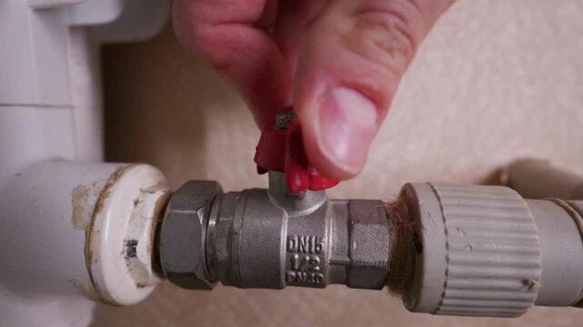Male hand turns a red butterfly handle on a metal ball valve connected to a home heating pipe. This manual action regulates the water flow or heat supply within a residential plumbing system.
