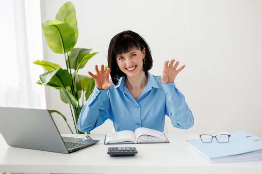 Happy female professional at a tidy home office desk with laptop, notebook and calculator, making a playful gesture during a video call conveys friendliness, confidence and approachable remote work