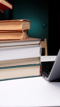 Stack of various books with a person holding another book, next to an open laptop on a white desk, representing education, research, and hybrid learning