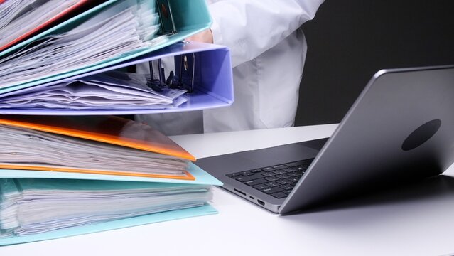 Professional person organizing many stacked binders full of files and important paperwork at a clean white desk, working with a laptop to manage and store documentation for bureaucracy and research