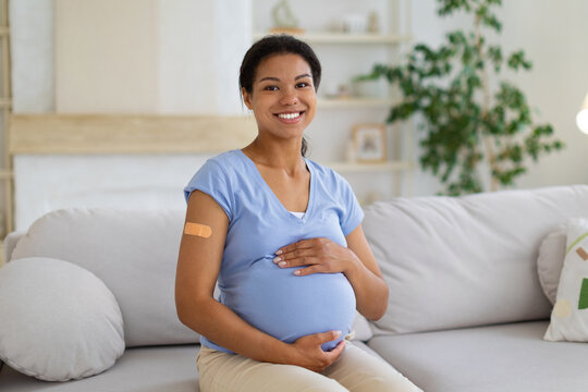 Smiling Black pregnant woman with vaccination bandage on her upper arm gently holds her belly while seated at home, promoting maternal health, protection, and wellness