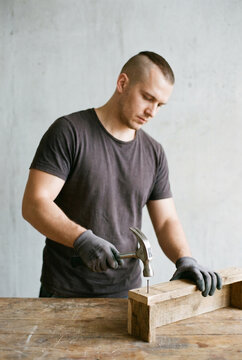 A man with a shaved undercut wearing a dark t shirt and gray work gloves hammers a nail into a wooden beam on a workbench
