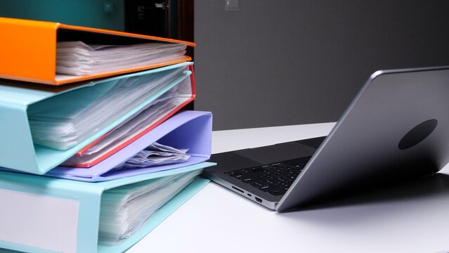 Stacked colorful binders containing documents and paperwork standing next to a modern laptop on a white desk