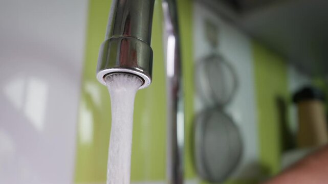 A chrome kitchen faucet pours a steady stream of clear water from its spout in a domestic setting. The blurred background shows a modern kitchen area with green wall tiles and hanging utensils.