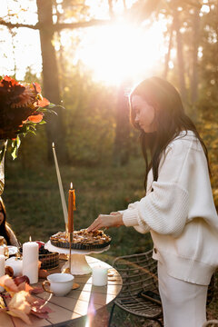 A woman in a cozy sweater serves a homemade pie at an outdoor candlelit table surrounded by autumn foliage and warm golden-hour sunlight, creating an intimate, peaceful seasonal celebration scene.
