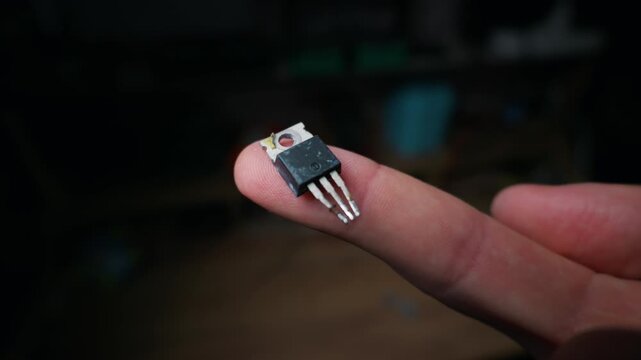 Small electronic transistor with three pins and a metal heat sink rests on a person finger tip against a dark background. Concept of microchip repair, hardware engineering, and circuit design.