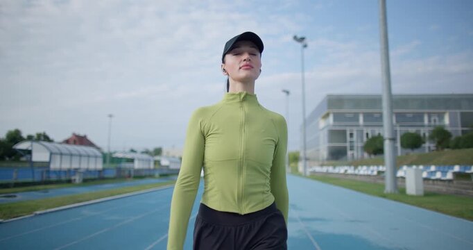 A young beautiful girl in sportswear and a cap with a skipping rope in her hands walks along the stadium's running track to train and exercise on a summer day