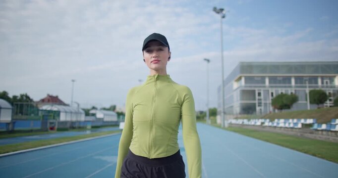 A young beautiful girl in sportswear and a cap with a skipping rope in her hands walks along the stadium's running track to train and exercise on a summer day