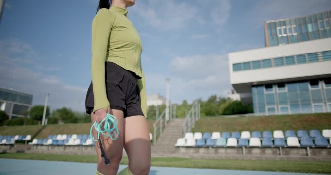 A young beautiful girl in sportswear and a cap with a skipping rope in her hands walks along the stadium's running track to train and exercise on a summer day