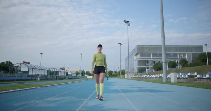 A young beautiful girl in sportswear and a cap with a skipping rope in her hands walks along the stadium's running track to train and exercise on a summer day