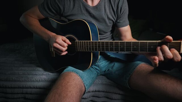 A man sits on a grey blanket while playing a blue acoustic guitar with a capo. Concept of creative expression, musical hobby, learning to play instrument, and relaxing indoor leisure.
