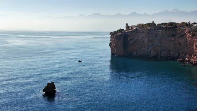 Small fishing boat drifts near rocky cliffs with distant mountains along the antalya coastline.