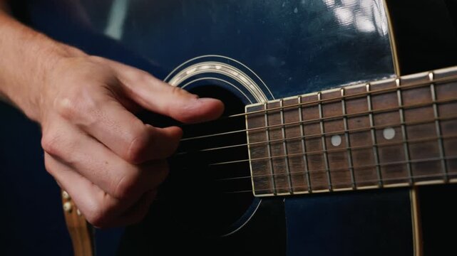 A close-up view of a musician fingers playing a dark blue acoustic guitar. The guitarist carefully plucks the steel strings while holding chords on the dark wooden fretboard.