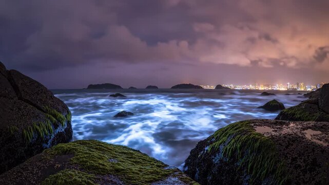 Bioluminescent waves flowing over rocky shoreline at night with city lights in background for nature and travel content