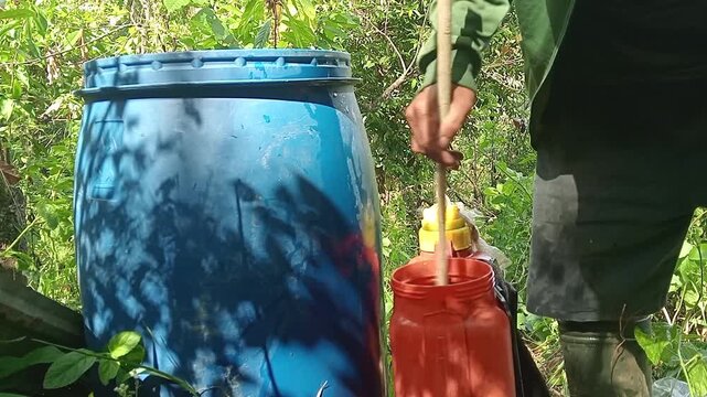 Farmer scooping water from blue barrel drum into sprayer tank outdoor
