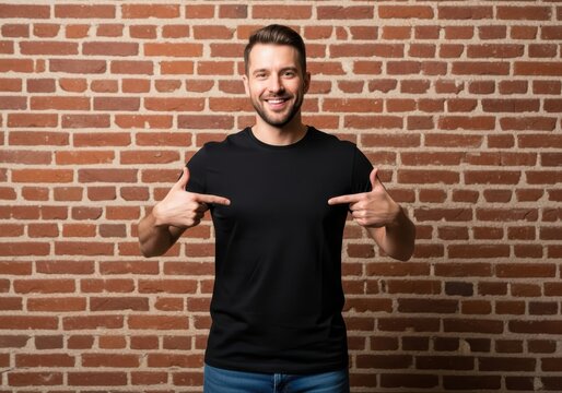 Smiling Young Man Pointing at Blank Black T-shirt Mockup Against Red Brick Wall Background for Apparel Design Presentation
