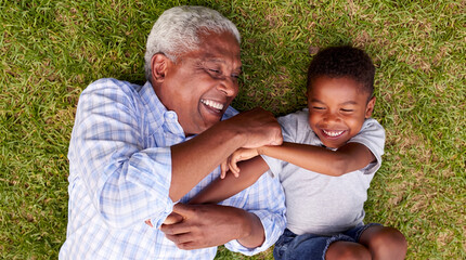 Fototapeta premium A cheerful grandfather and his young grandson lying on the green grass, playing and laughing together in a beautiful park.