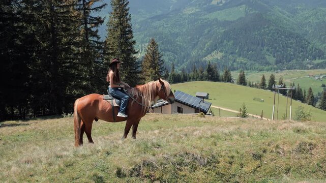 Female rider pauses on horseback in a broad highland meadow landscape, Wide view shows forested mountain slopes and rural buildings in daylight