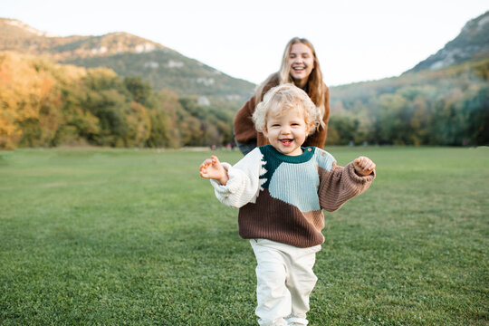 Smiling little baby girl 1-2 year old running from her mother in green grass lawn and mountain landscape at background. Motherhood.