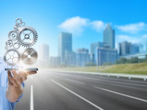 A hand holds a smartphone beneath silver gears, symbolizing business automation along a road toward the city.
