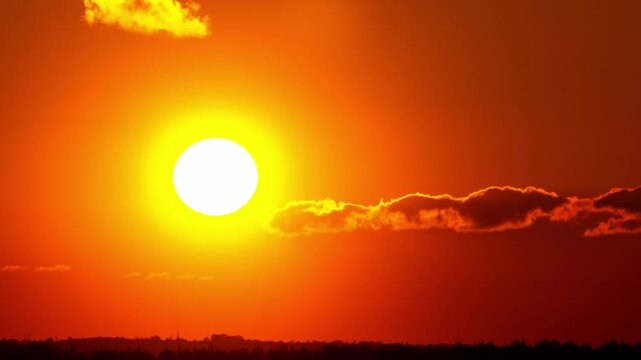 Timelapse of a large bright sun descending through a fiery red and orange sky. A few wispy clouds drift past as it sets behind a dark silhouetted treeline.