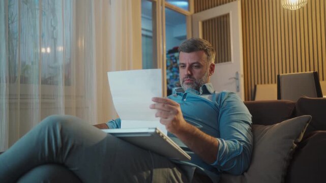 View of a man sitting on a sofa and looking at household bills and paperwork, desperately sighing