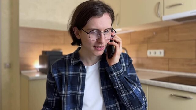 A young man talks on the phone while sitting at a kitchen table. He holds a piece of paper in his hand, focused on the conversation and his notes.