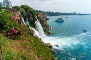 Fototapeta premium Antalya, Turkey. Lower Duden Waterfall plunging from a flower-covered cliff into the turquoise Mediterranean, with a sightseeing boat below and the modern city skyline beyond